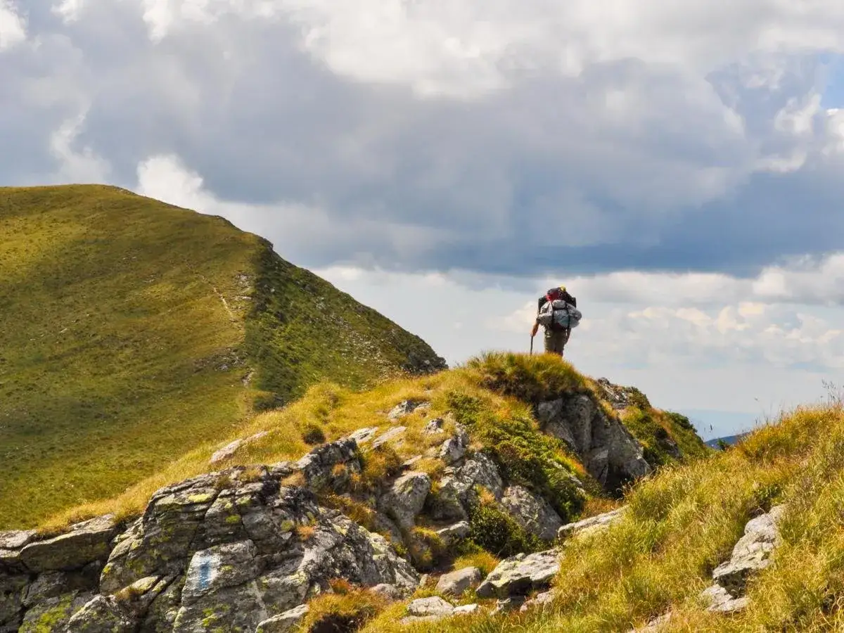 Ein Wanderer mit Rucksack und Stock steht auf einem felsigen Grat. Im Hintergrund erstreckt sich ein gr&uuml;ner Berghang. Ein tolles Erlebnis beim Karpaten wandern.