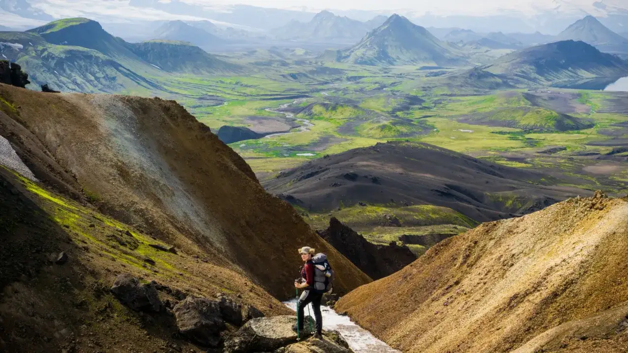 Eine Wanderin mit Rucksack und Stöcken steht auf einem Felsen. Die weite Landschaft mit grünen Hügeln und Bergen zeigt die wahre trekking bedeutung.