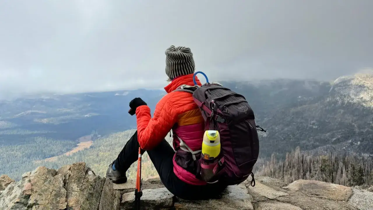 Nach mehreren Tagen im Gebirge wandern, sitzt eine Person mit Rucksack und Wanderstöcken auf einem Felsen und blickt in die nebelverhangene Berglandschaft.