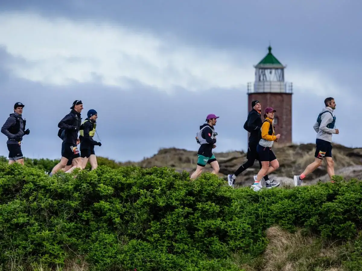 Ultramarathonl&auml;ufer laufen an einem Leuchtturm vorbei durch eine gr&uuml;ne, h&uuml;gelige Landschaft.