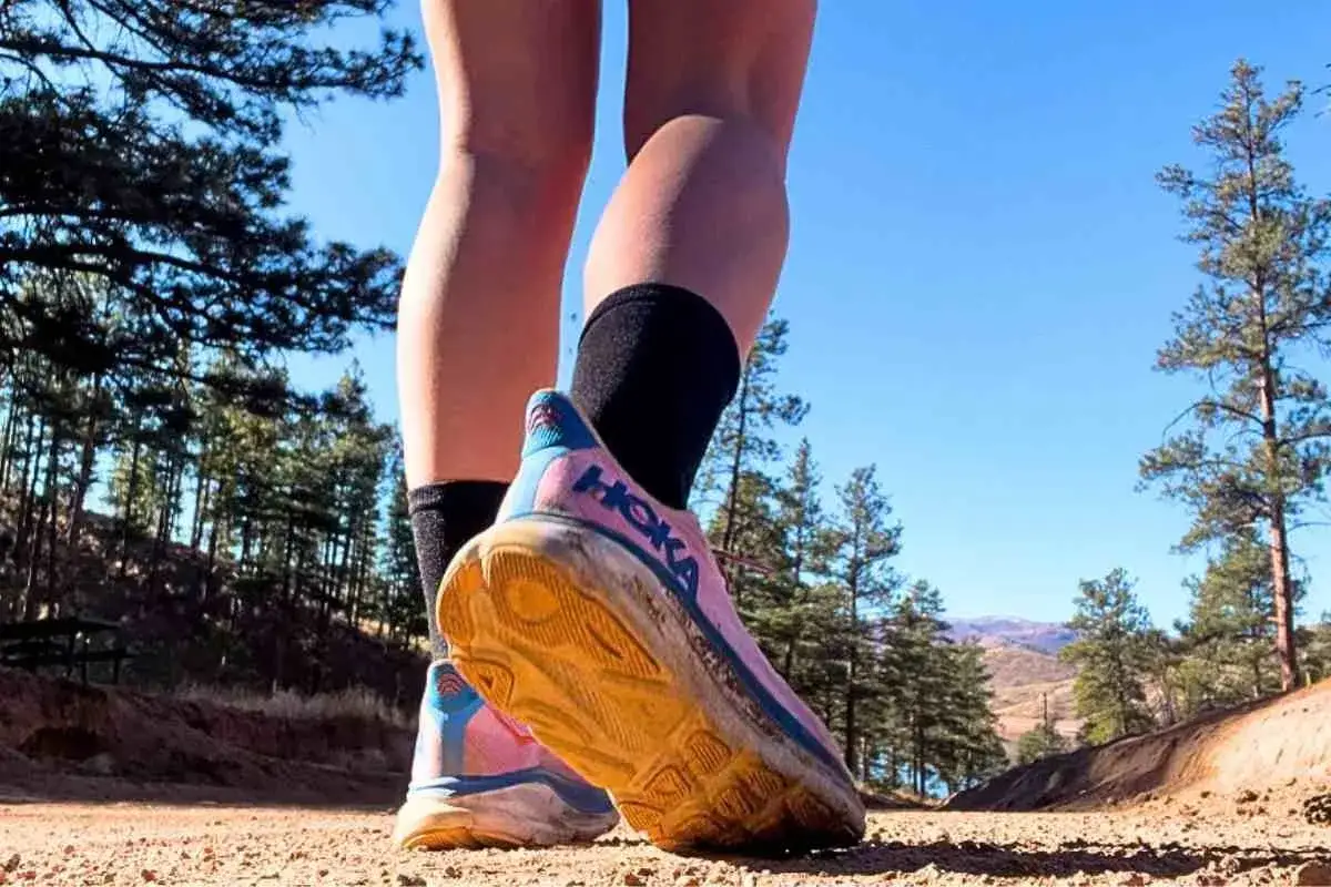 Läuferin im Hoka Clifton 9 Test auf staubigem Trail. Die Schuhe sind pink mit blauen Akzenten und zeigen deutliche Gebrauchsspuren.