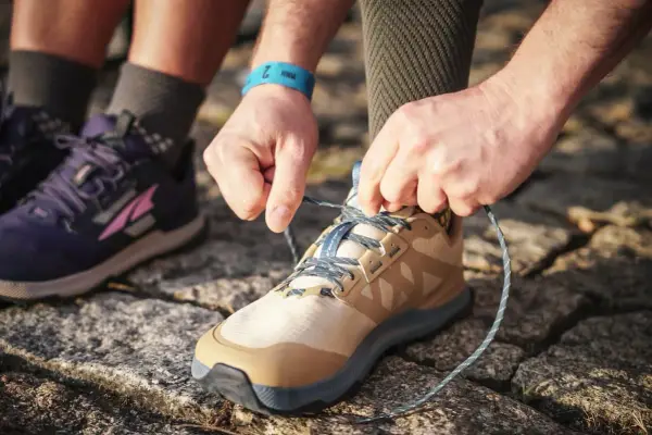 Hände binden Schnürsenkel eines beigefarbenen Schuhs, bereit für den Trail. Daneben ein lila Schuh, der verschiedene wanderschuhe kategorien zeigt.