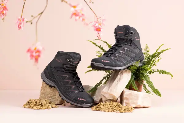Zwei schwarze Wanderschuhe von LOWA, auf Steinen und Kies arrangiert, mit zarten rosa Blüten und Farn im Hintergrund.