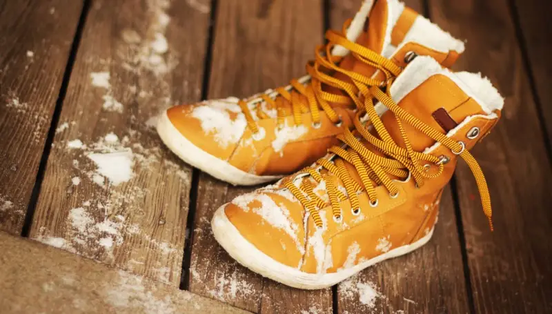 Warme, gefütterte Stiefel mit gelben Schnürsenkeln liegen auf einer Holzplanke, bedeckt mit etwas Schnee. Sie warten darauf, dass die **Schuhe trocknen**.