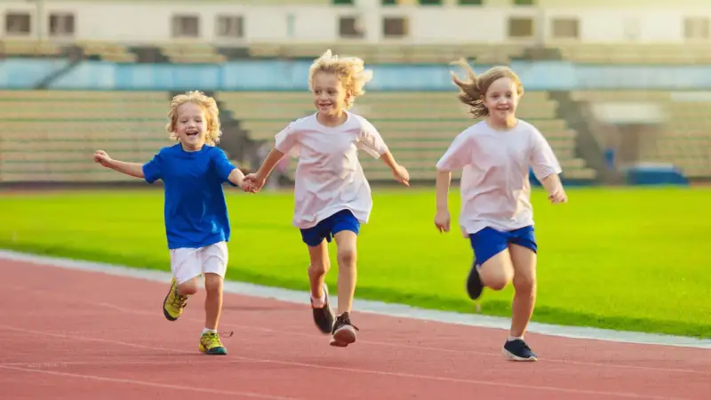 Drei lachende Kinder beim Lauftraining auf einer roten Laufbahn. Sie rennen nebeneinander, die Arme ausgestreckt.